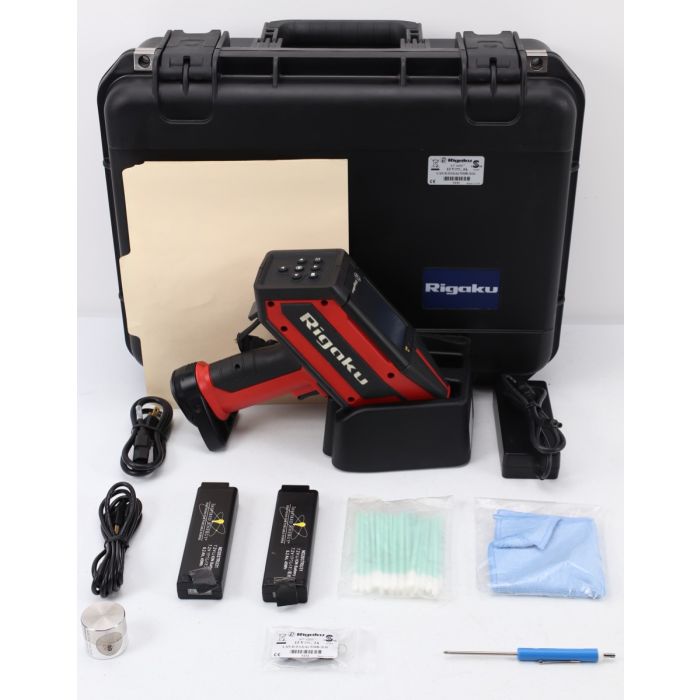 The image shows a Rigaku KT-100S X-ray Fluorescence Analyzer, a compact, white-colored device with various buttons and a display screen on the front. The device is placed on a gray-colored table or platform, and there is a black-colored wire attached to i