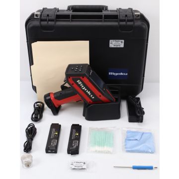 The image shows a Rigaku KT-100S X-ray Fluorescence Analyzer, a compact, white-colored device with various buttons and a display screen on the front. The device is placed on a gray-colored table or platform, and there is a black-colored wire attached to i